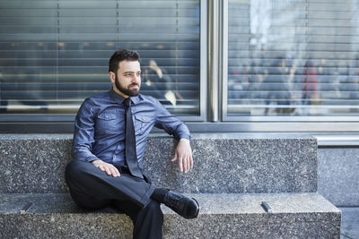 Full length of thoughtful businessman sitting on seat against window in office