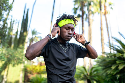 Young african american male athlete in sportswear listening to music with his headphones keeping hands on the headphones and closed eyes while standing in green tropical park