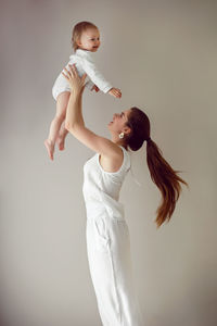 Mom throws her daughter child in her arms stands next to window in white clothes at home in winter