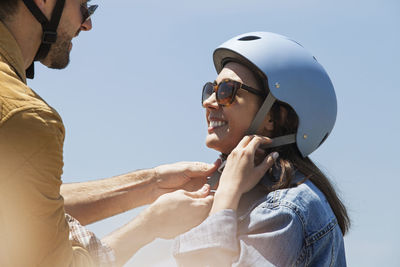 Man assisting woman in wearing bicycle helmet against clear sky on sunny day