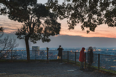 Rear view of people standing by railing against sea during sunset