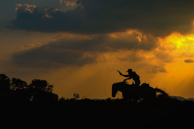 Silhouette cowboy aiming from gun while riding horse on land during sunset