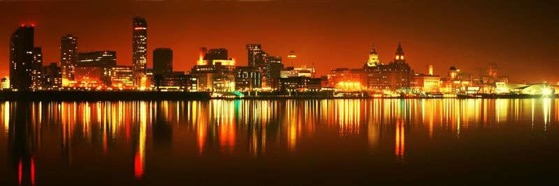 Illuminated cityscape by lake against sky at night