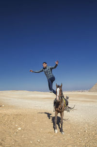 Full length of mid adult man jumping from horse on sand against clear blue sky during sunny day