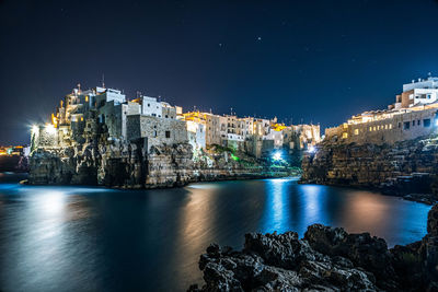 Illuminated buildings by sea against sky at night