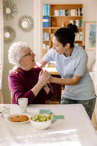 Smiling retired senior woman holding hands of young female volunteer while talking in nursing home