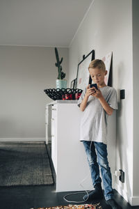 Full length of boy charging smart phone while standing by wall in room at home