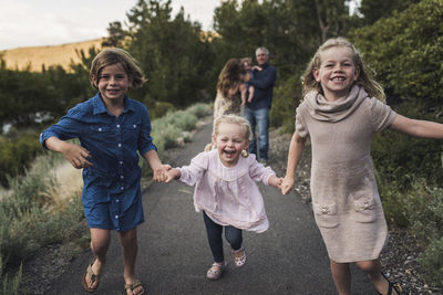 Happy sisters holding hands while running on road against parents in forest