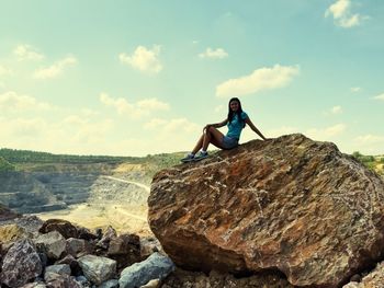 Young woman sitting on rock against sky