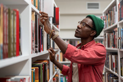 Thoughtful african american student man in glasses choosing book in college library or bookstore