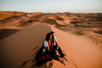 Man sitting on sand dune in desert against sky