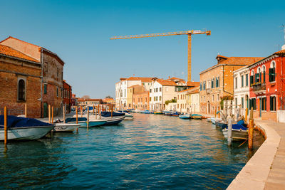 Murano canal with colorful houses, moored boats and cranes in the background