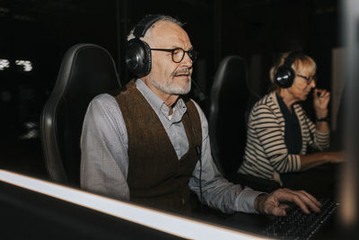 Senior man wearing headset playing video game on computer by female friend at gaming center