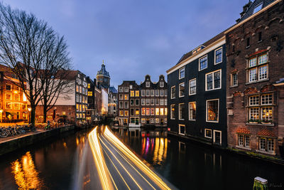 Light trails on river amidst illuminated buildings in city against sky at dusk