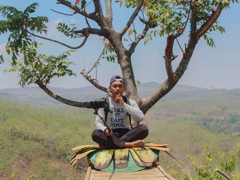 Young man taking selfie while sitting on tree against sky