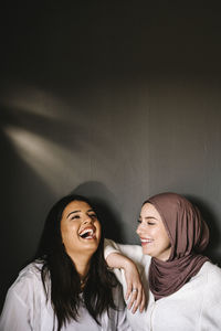 Happy young woman wearing hijab looking at friend laughing against gray background