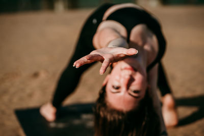 Tranquil female standing on mat in camatkarasana and reaching out towards camera while doing yoga on beach in summer