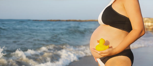 Midsection of pregnant woman with rubber duck standing at beach