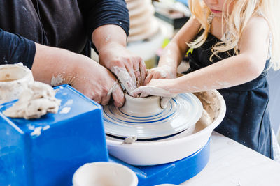 Crop unrecognizable female in black casual outfit standing near pottery wheel while shaping clay pot with little girl in light workshop