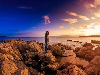 Man standing on rock by sea against sky during sunset