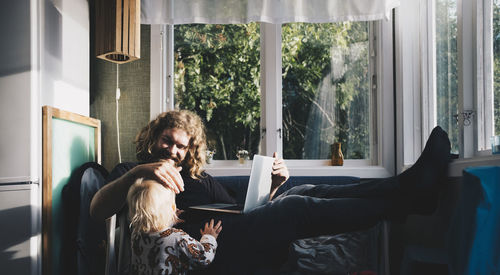 Father holding laptop while looking at girl by window