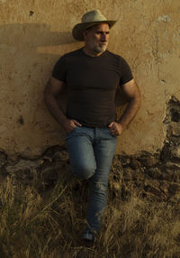 Portrait of adult man in cowboy hat and jeans against abandoned building during sunset