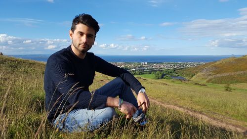 Portrait of young man sitting on field by sea against sky