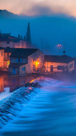 Illuminated houses by river against sky at sunset