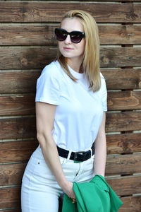 Portrait of young woman wearing sunglasses while standing against wall