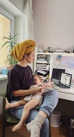 Woman wearing yellow towel on her head, blue training pants working from home and breastfeeding baby