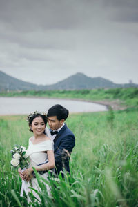 Married couple embracing amidst grass against lake