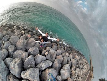 Full length of man with daughters standing on stones at sea shore against cloudy sky