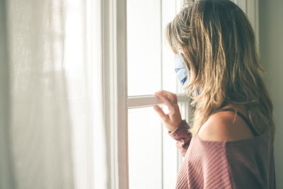 Woman looking outside the window with surgical mask during coronavirus lockdown. covid-19 people