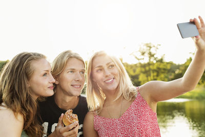 Happy friends taking selfie while having breakfast by lake against clear sky