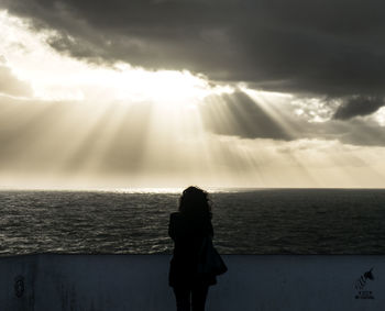 Silhouette woman standing on sea shore against sunbeam streaming through cloudy sky during sunset