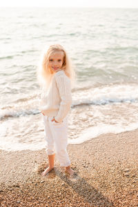 Cute stylish kid girl 5-6 year old wear white knit sweaterand pants rest over sea shore background. 