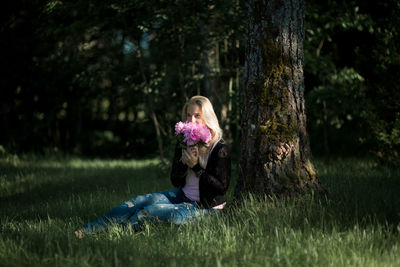 Young woman smelling flowers while sitting on grassy field in forest