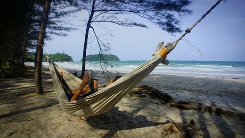 Woman reading book while relaxing on hammock at beach against sky