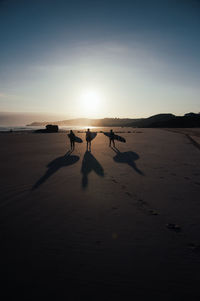 Friends carrying surfboard while walking at beach against clear sky during sunset