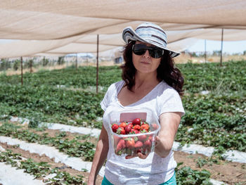 Portrait of woman holding strawberries in container while standing at farm during sunny day
