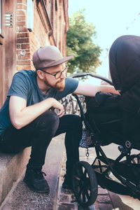 Full length of mid adult man peeking in baby carriage while sitting on steps outdoors