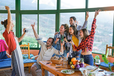 Happy friends holding wineglasses while standing against windows at home