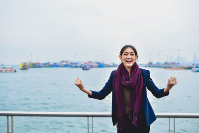 Cheerful young woman clenching fists while standing against sea