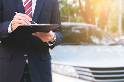 Midsection of salesman holding clipboard while standing against car