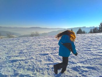 Rear view of girl running on snow covered field against blue sky