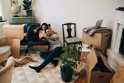 Smiling couple video calling through mobile phone while sitting in living room during relocation