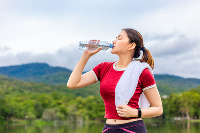 Young woman drinking glass while standing against water