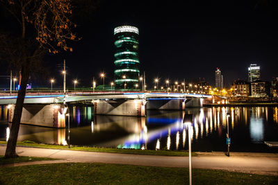 Illuminated bridge over river by buildings against sky at night