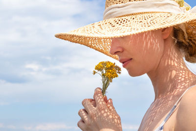 Close-up of woman wearing hat smelling flower against sky