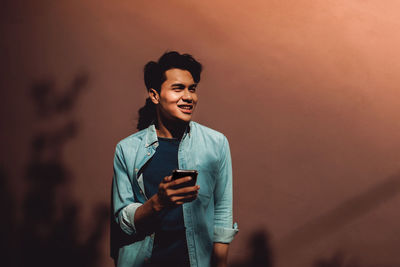 Smiling young man using phone while standing against wall